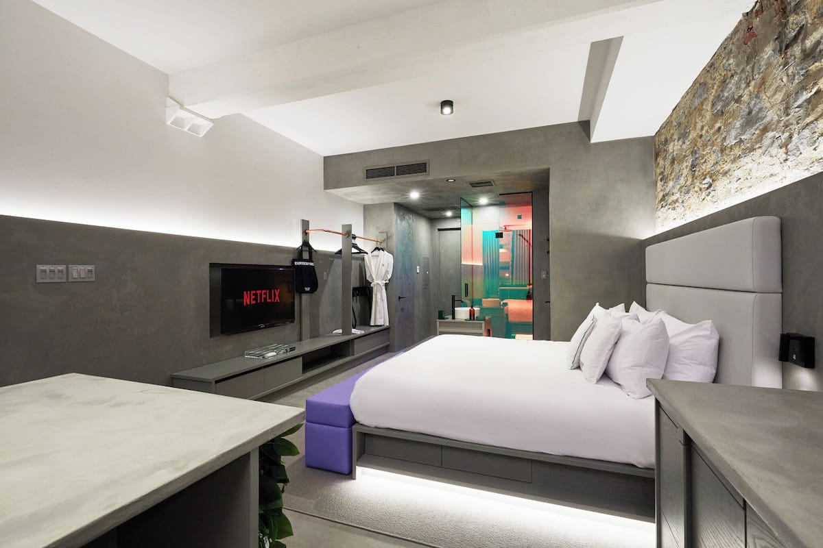 Una habitación de hotel moderna con una cama grande, televisión, zona de ejercicios y un baño visible a través de una puerta abierta.