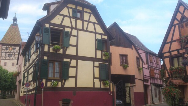 A traditional half-timbered building with a red base and white upper sections, featuring multiple windows with shutters and hanging flower baskets. - GITES LE BRETZEL (Bergheim)