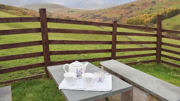 Una mesa con un calentador de té y tazas, con vistas a un paisaje cubierto de hierba y una valla de madera.