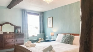 Quarto com cama de madeira, cômoda, cadeira, janela com cortinas, abajur e quadro na parede.