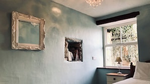 Quarto com cama, mesa de cabeceira, luminária, janela com vista, espelho fixado na parede e detalhe de parede em pedra.