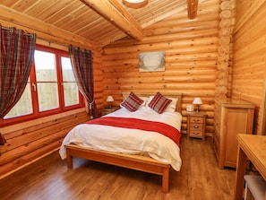 Een slaapkamer in een blokhut met een houten bed, twee nachtkastjes, een kaptafel en een raam met ruitjesgordijnen.