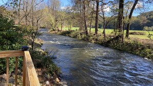 A wooden bridge over a flowing river with trees and a grassy field in the background. - Tiny Toot - “On Deep Creek” Bryson City, NC  (Bryson City)