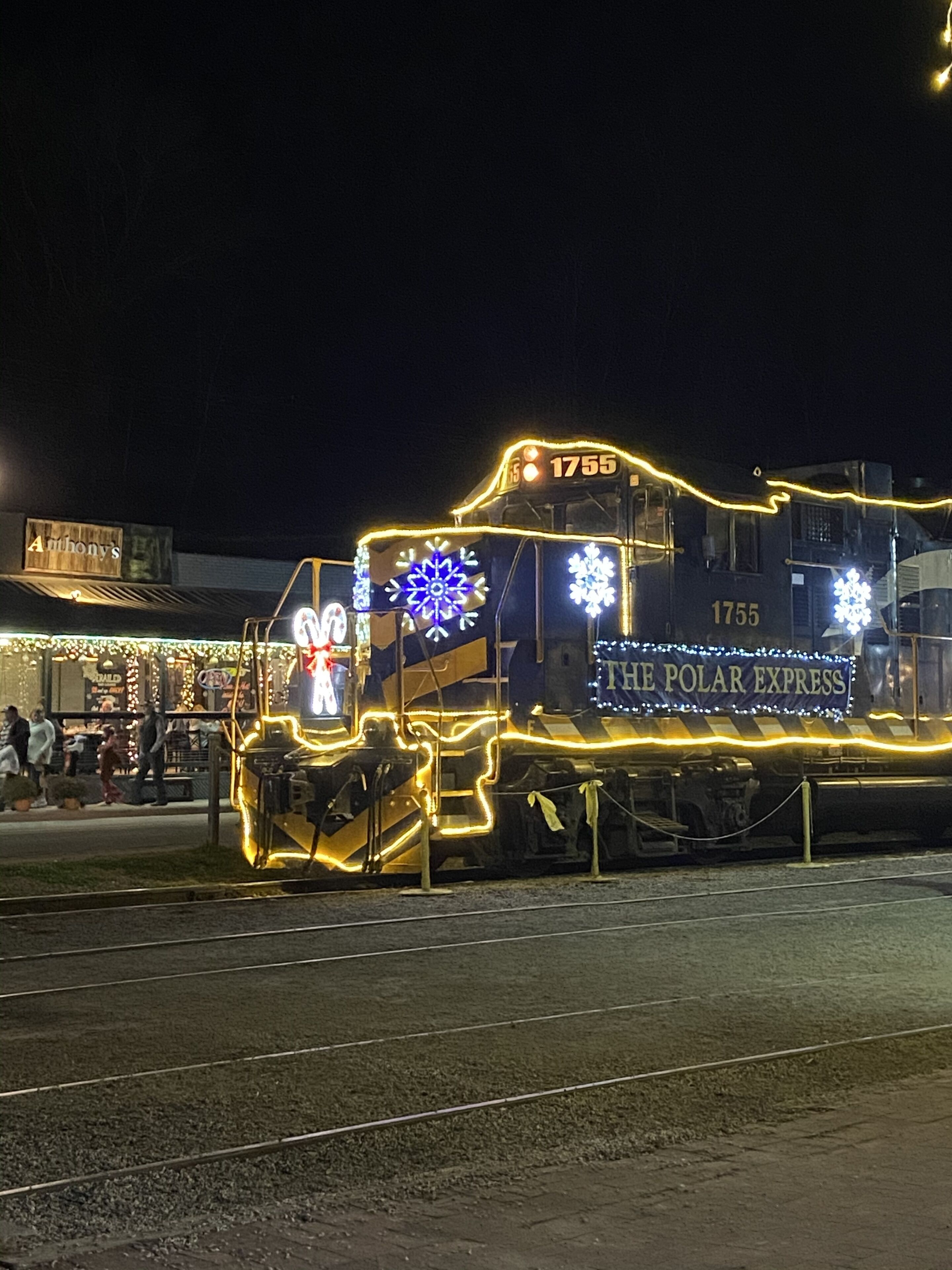 A stationary train with the number 1755 and the name "THE POLAR EXPRESS" on its side, decorated with lights and snowflake patterns.