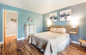 A bedroom with a bed, dresser, bedside table, and artwork on the wall.