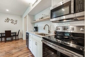 A modern kitchen with a stainless steel stove, microwave, and built-in oven. There is a wooden dining table with chairs and a white cabinet with a sink.