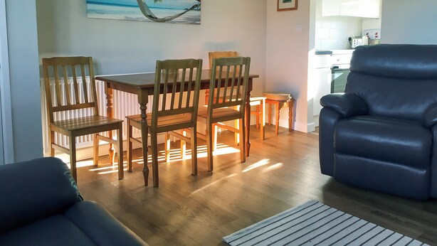 Un coin repas avec des chaises et une table en bois, un fauteuil bleu et un tableau représentant une scène de plage accroché au mur.