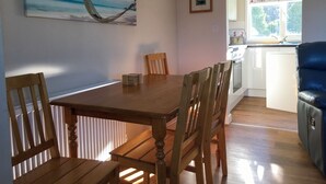 Un coin repas avec une table et des chaises en bois, un tableau représentant une scène de plage, une photo encadrée accrochée au mur et une vue sur la cuisine par une porte ouverte.