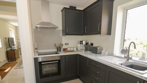 A modern kitchen with dark cabinets, a built-in oven, a stainless steel range hood, and a window with a view of greenery. - Garden Cottage (Worcester)
