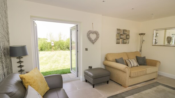 A living room with a grey sofa, beige sofa, and a grey ottoman. There is a large window with a view of a garden. - Garden Cottage (Worcester)