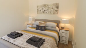 A neatly made bed with a quilt, two bedside lamps, a nightstand with a lamp, and a wall-mounted artwork above the bed. - Garden Cottage (Worcester)