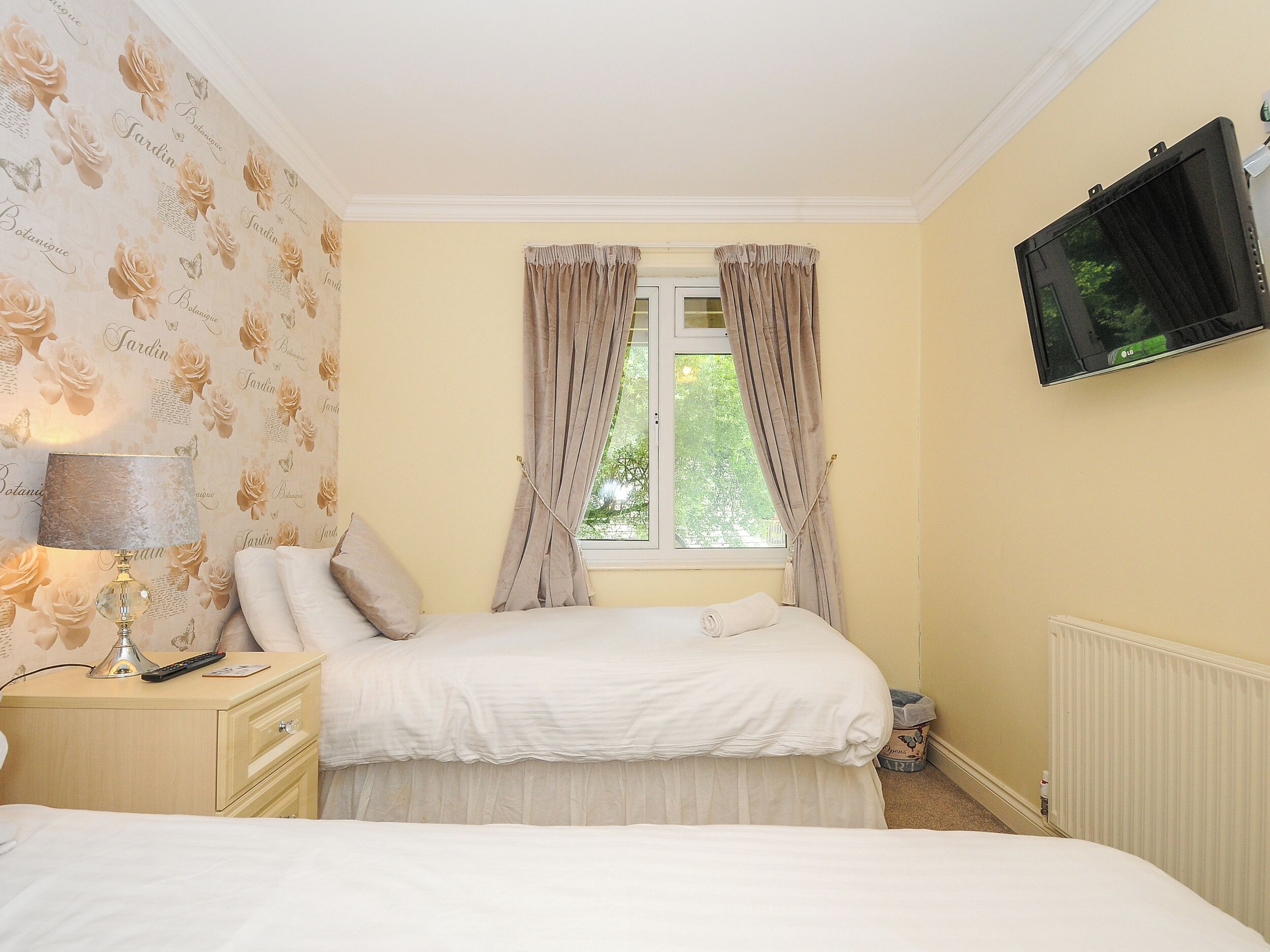 A bedroom with a bed, a nightstand, a lamp, a TV, and a window with curtains.