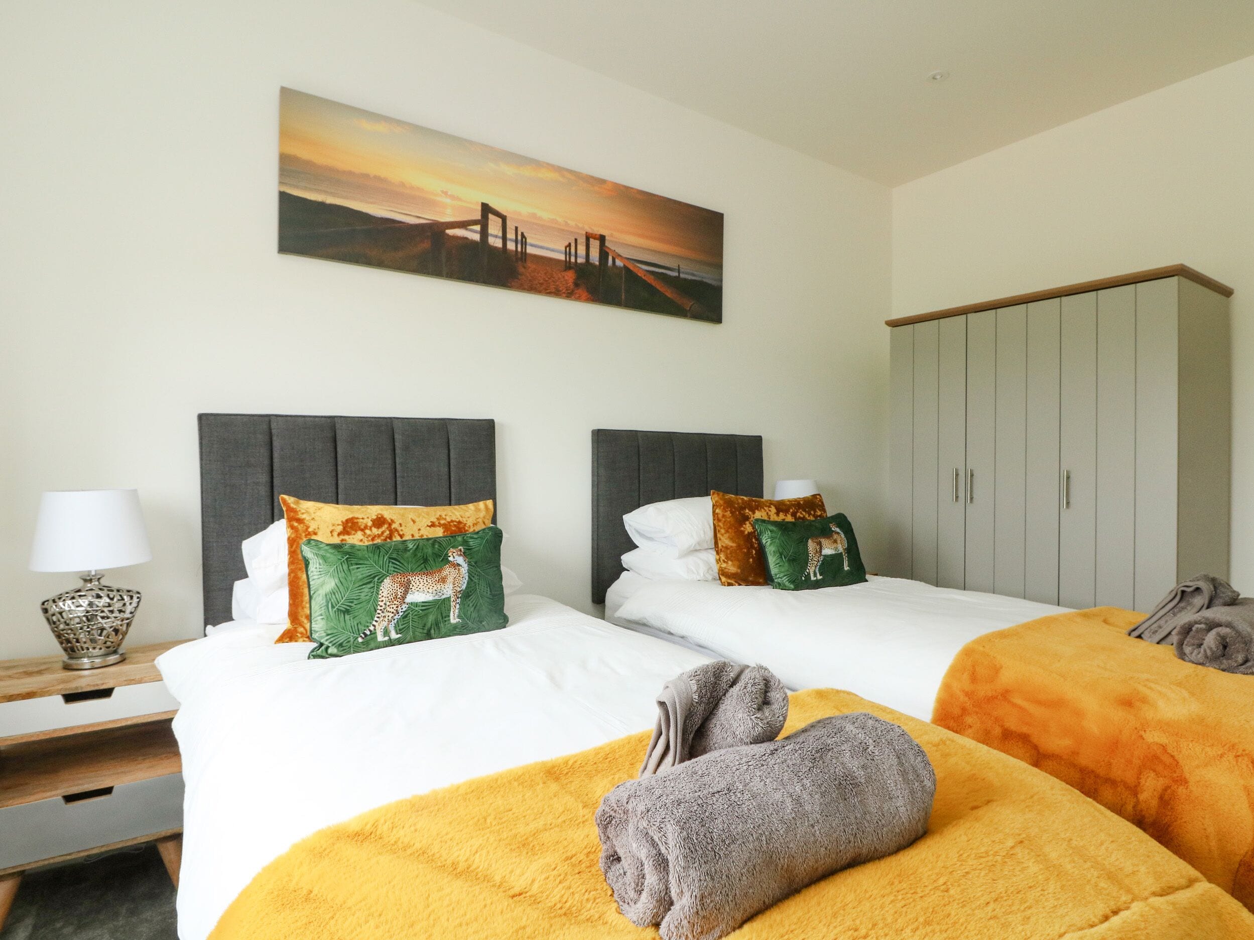 A hotel room with two beds, each with a grey headboard, white bedding, and decorative pillows. There is a painting above the beds, a wardrobe, and a bedside table with a lamp.