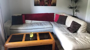 A living room with a beige sectional sofa, a glass-top coffee table, a red throw blanket, and a painting of two figures on the wall. - Immaculate 3-bed Apartment in Kilkenny (Kilkenny)