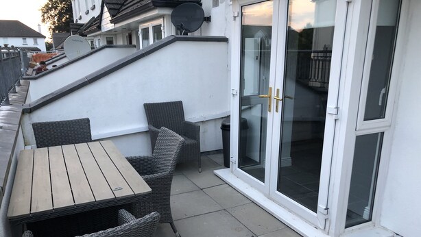 A patio with a wooden table and wicker chairs, a glass door, and a white wall. - Immaculate 3-bed Apartment in Kilkenny (Kilkenny)