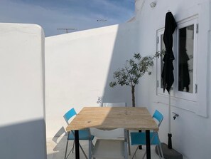 Un petit espace extérieur aménagé pour prendre les repas, avec une table en bois et des chaises bleues, une plante en pot et un mur blanc.