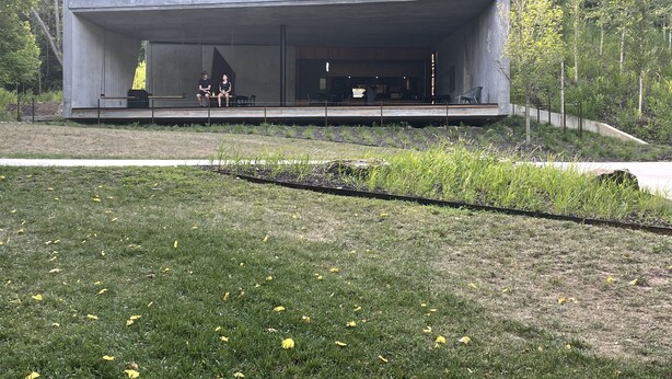A modern building with a flat roof, large windows, and a flat facade. There are two people seated on a bench outside.