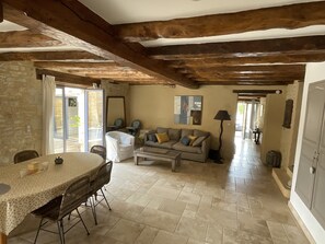 A spacious living area with a stone wall, wooden beams, a dining table with chairs, a sofa with cushions, and a floor lamp.