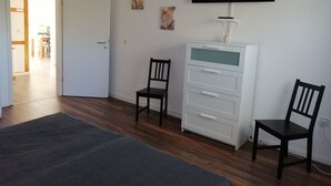 A room with a flat-screen TV mounted on the wall, a white cabinet with drawers, two wooden chairs, and a grey rug on the floor.