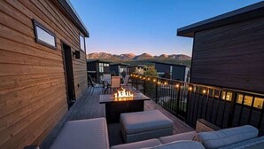 A wooden deck with outdoor seating, a fire feature, and mountain views. - Bring Fido | Pool & Hot Tub | Amazing Location (Whitefish)