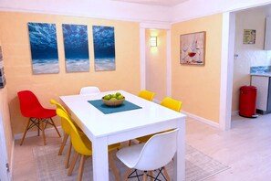 A dining area with a white table, yellow and red chairs, a bowl of fruit, and three ocean-themed paintings on the wall.