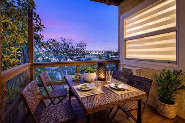A balcony with wicker furniture, a table set for two, and a view of a cityscape at dusk.