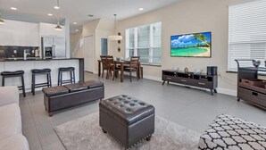 A modern kitchen with bar stools, a dining area with a table and chairs, a living room with a sofa and ottoman, and a large window with blinds. - 3 bed Modern Town Home On a Luxury Development Close to Disney (Clermont)