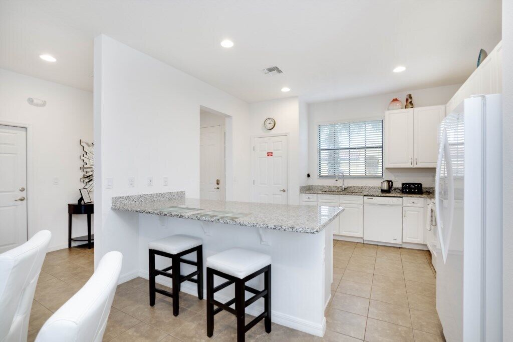 A modern kitchen with white cabinetry, a granite island with bar stools, and a window with blinds.
