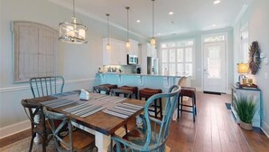 Uma sala de jantar espaçosa com uma mesa e cadeiras de madeira, uma cozinha ao fundo e iluminação pendente.