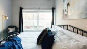 Un dormitorio con cama, ventana, lámpara de pie y un cuadro en la pared.