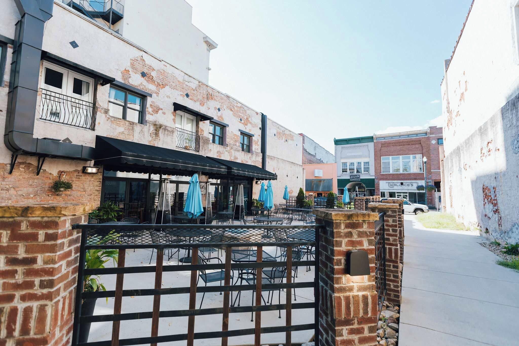 A street view with a building featuring a brick facade, a metal gate, and outdoor seating with umbrellas.