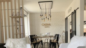 Een moderne eethoek met een bakstenen muur, een houten scheidingswand met de decoratie 'hello' en 'CAENA AMAL', een ronde tafel met stoelen en een surfboard aan de muur.