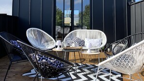 Een modern buitenzitgedeelte met witte rieten stoelen en een kleine tafel, een zwart metalen dak en een groot raam met uitzicht op een landschap.