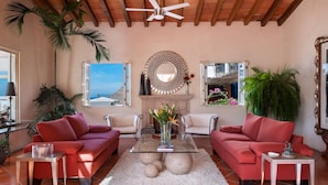 Sala de estar con dos sofás rojos, una mesa de centro de vidrio y una chimenea. Hay plantas y un espejo grande en las paredes.