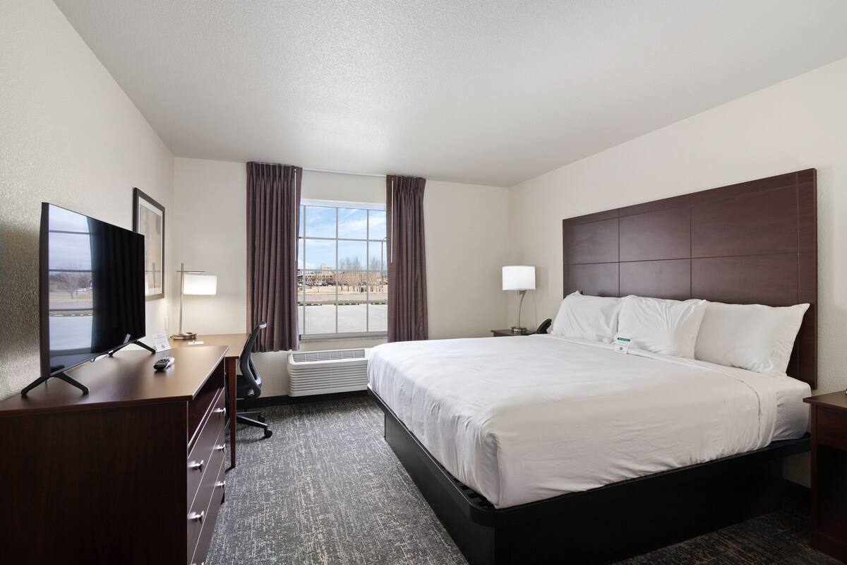 A hotel room with a large bed, a desk with a computer, a TV, and a window with curtains.