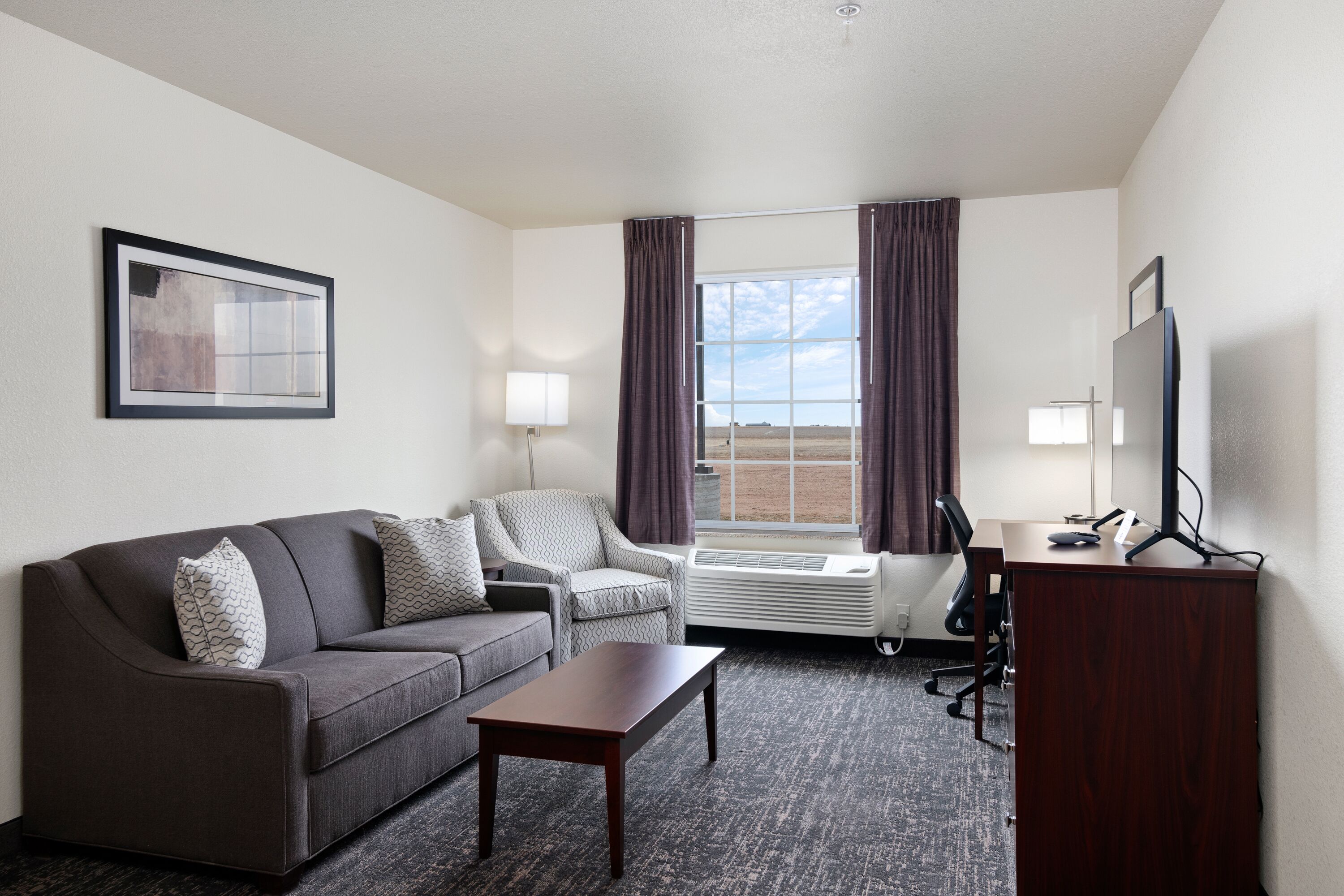 A hotel room with a gray sofa, a small table, a desk with a computer, a TV, and a window with curtains.