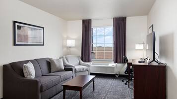 A hotel room with a gray sofa, a small table, a desk with a computer, a TV, and a window with curtains.