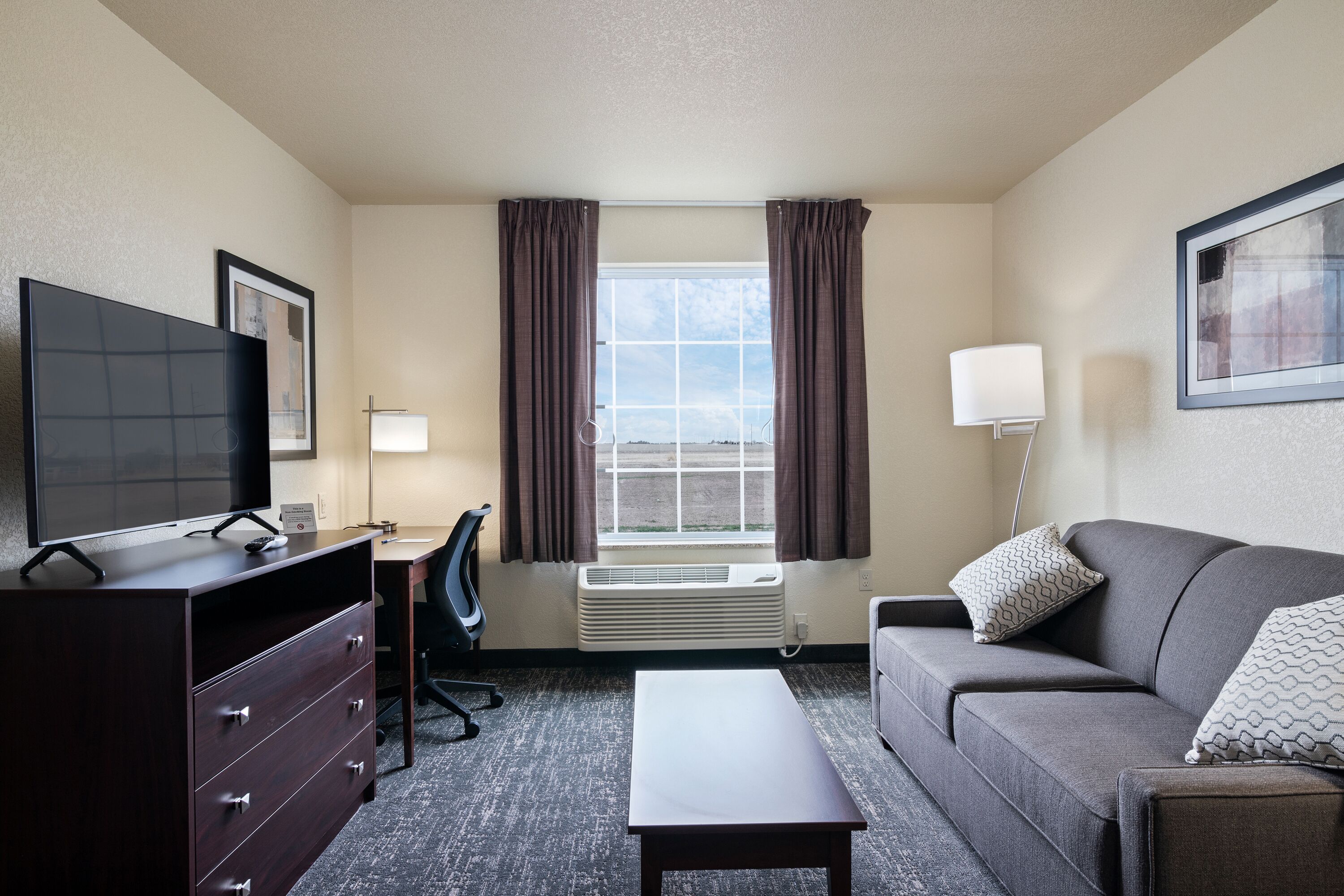 A hotel room with a TV, desk, chair, couch, and a window with curtains.