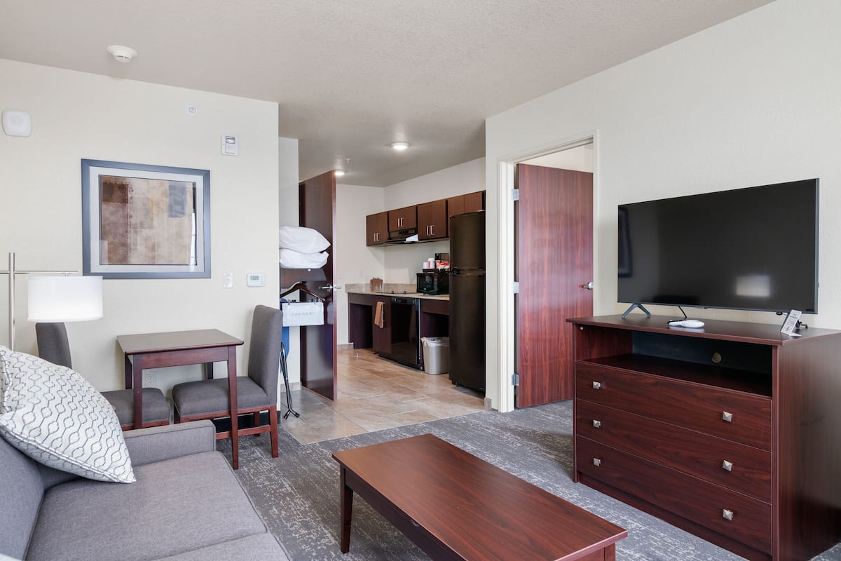 A modern living room with a sofa, coffee table, TV stand, and kitchen area with cabinets and appliances.