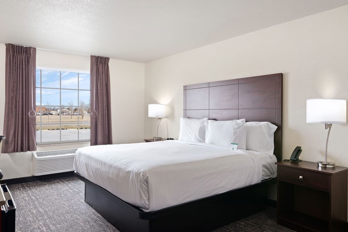 A hotel room with a large bed, two bedside lamps, a desk with a phone, a window with curtains, and a view of the outside.