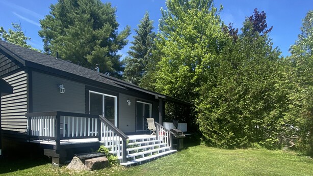 A single-story house with a front porch, a small deck, and a grassy yard. - 3 BEDROOM LAKE FRONT COTTAGE WITH SPECTACULAR LAKE VIEWS AND SERENE ATMOSPHERE (Utterson)