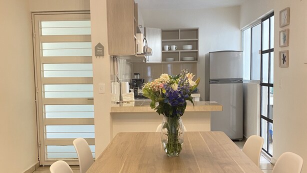 A dining area with a wooden table and white chairs, a vase with flowers, and a kitchen in the background. - LUNA • Calm 2BR 2BA Apartment • Fast WiFi • Spacious Stay (Puerto Vallarta)