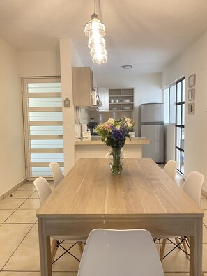 Un comedor con una mesa de madera y sillas blancas, un florero con flores, y una cocina al fondo.