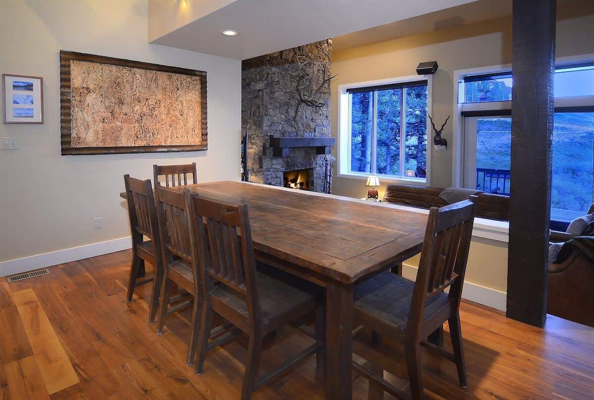 A dining area with a wooden table and chairs, a stone fireplace, and a large window with a view of trees.