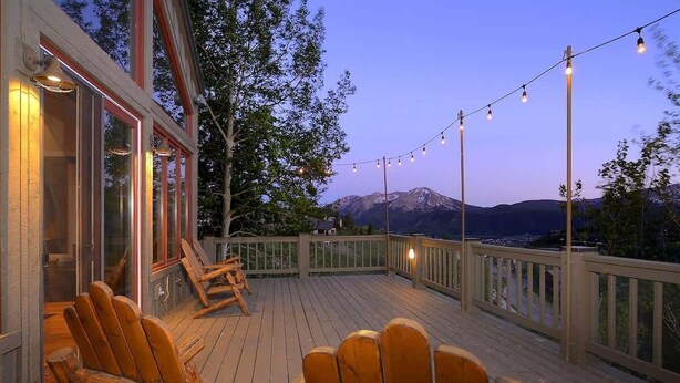Una terraza de madera con sillas Adirondack, luces colgantes y vistas a montañas.