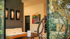 Una habitación moderna con un banco de madera, un sofá blanco y una silla de mimbre. Hay un cuadro abstracto colorido en la pared y una puerta con panel de vidrio esmerilado.