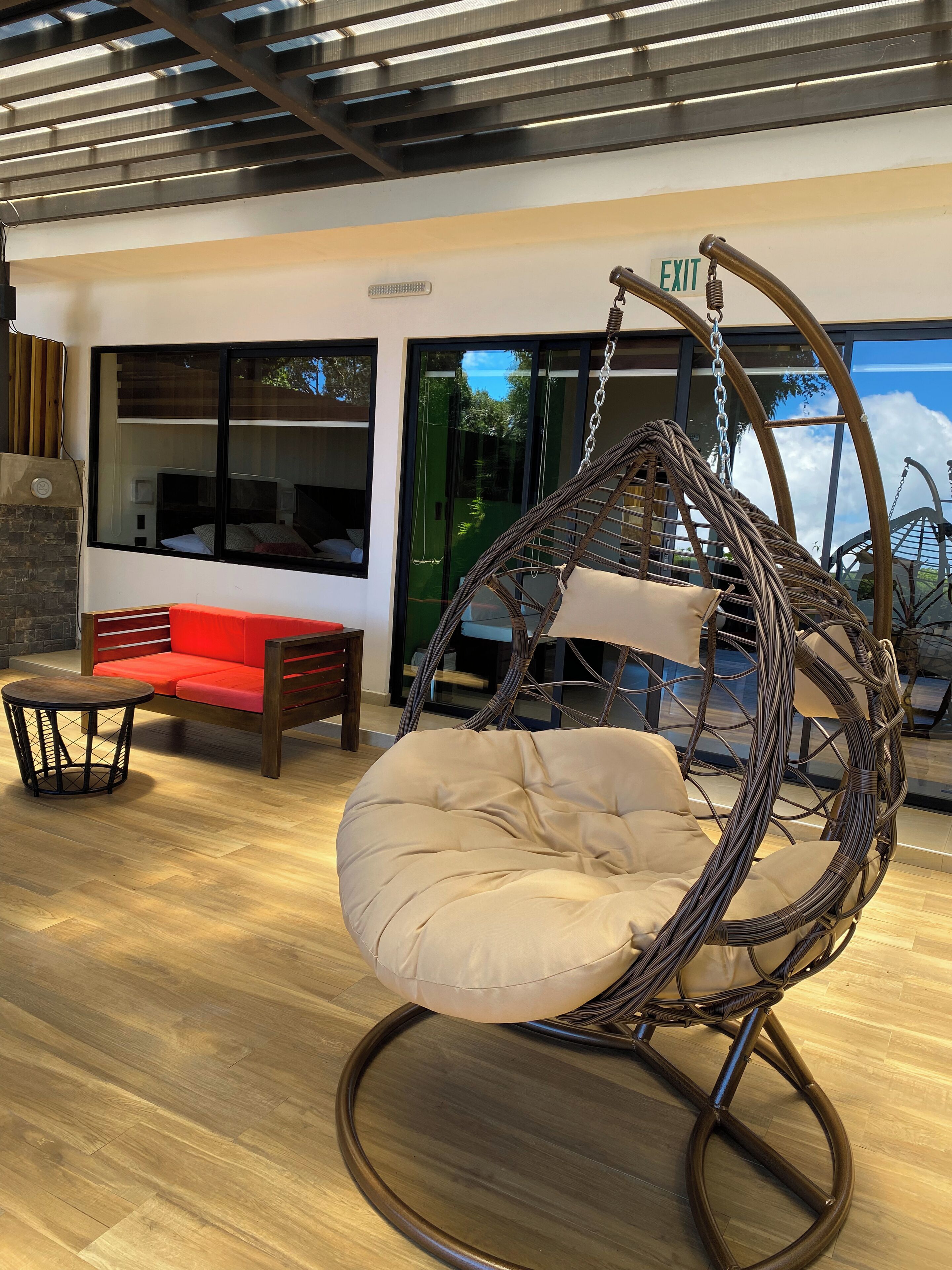 A wicker hanging chair with a cushion in a modern room with a wooden floor, a red sofa, and a glass door leading outside.