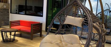 A wicker hanging chair with a cushion in a modern room with a wooden floor, a red sofa, and a glass door leading outside.