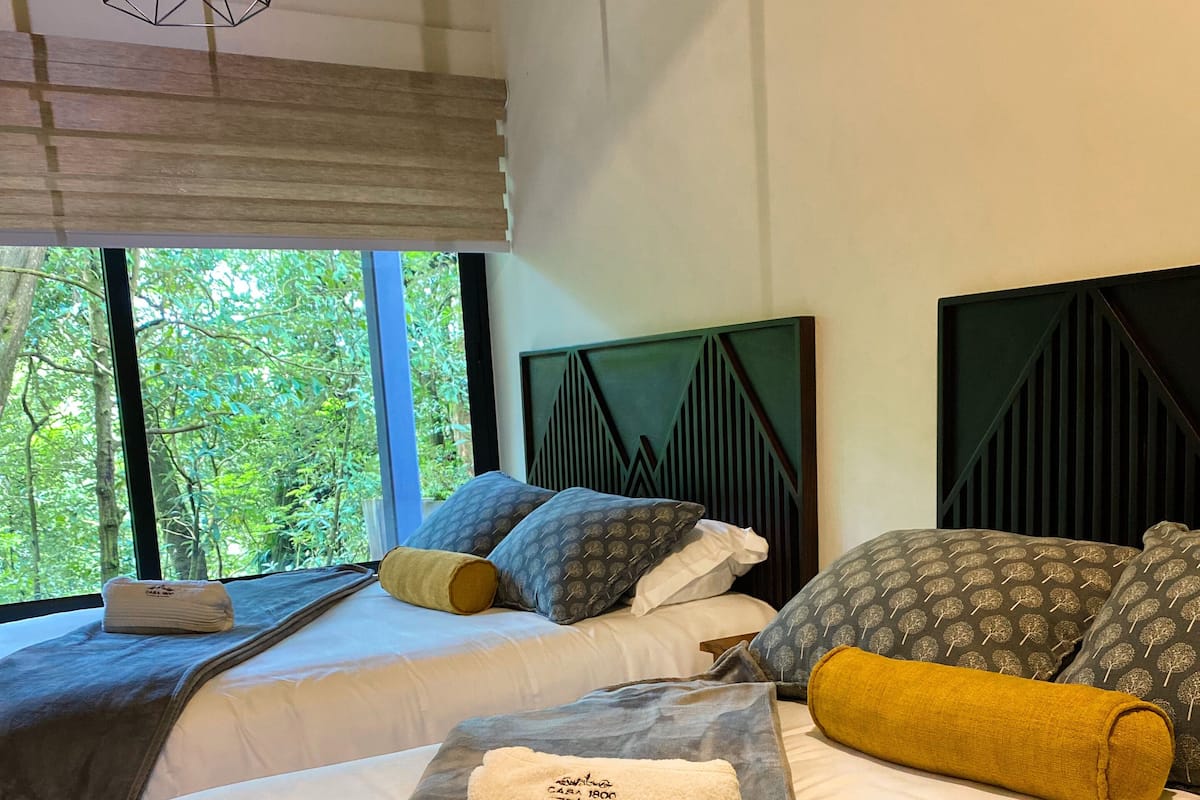 Habitación de hotel con dos camas, un ventanal con vistas a árboles y un cabecero de estampado geométrico.
