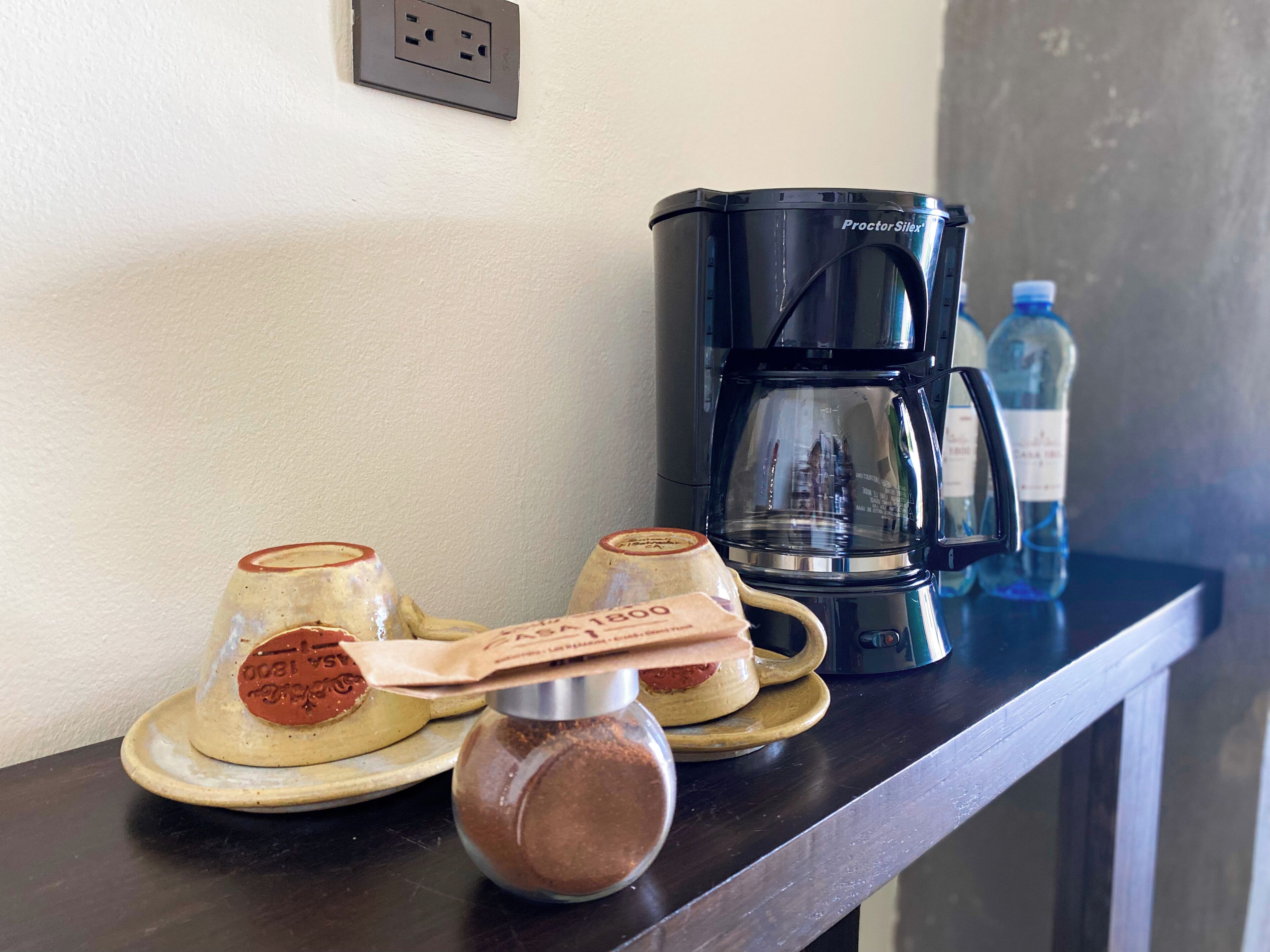 Uma cafeteira, duas chávenas e um frasco de especiarias numa bancada.